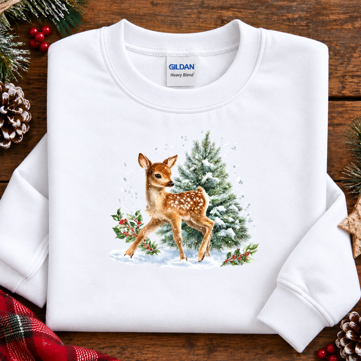 A folded white Gildan Heavy Blend sweatshirt on a rustic wooden background, decorated with pine branches, red berries, and a pinecone. The front of the sweatshirt features a detailed watercolor-style graphic of a brown spotted fawn standing next to a snowy green pine tree.