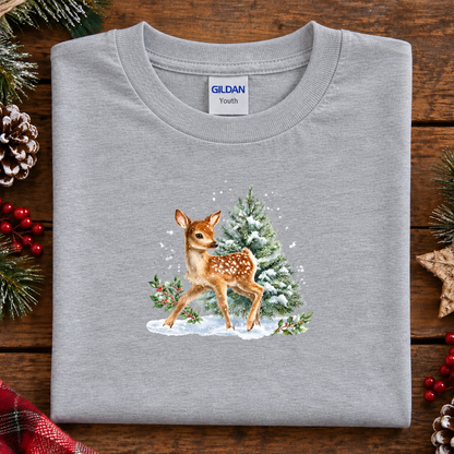 A folded sport gray Gildan Youth t-shirt with a watercolor-style graphic of a fawn and a Christmas tree, displayed on a rustic wooden background with holiday decorations.