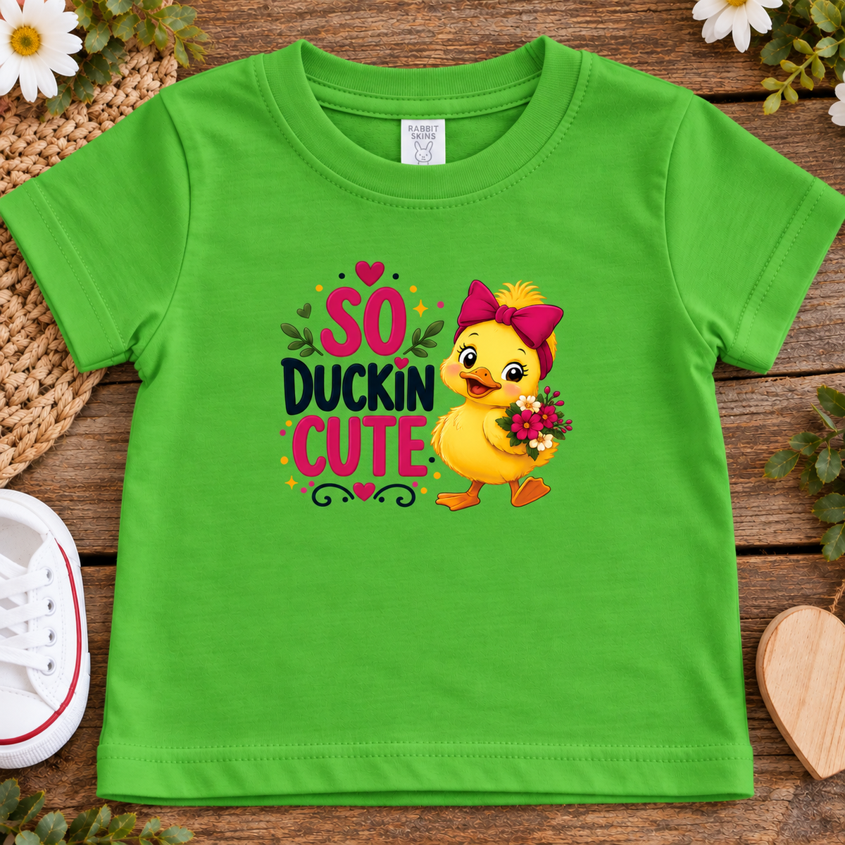 A apple green toddler t-shirt with a 'So Duckin Cute' graphic print, laid flat on a rustic wooden surface with decorative flowers, a woven mat, and a white sneaker.