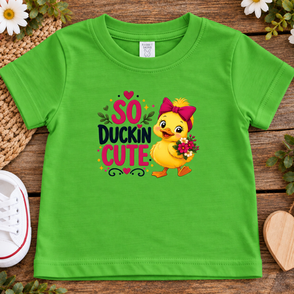 A apple green toddler t-shirt with a 'So Duckin Cute' graphic print, laid flat on a rustic wooden surface with decorative flowers, a woven mat, and a white sneaker.