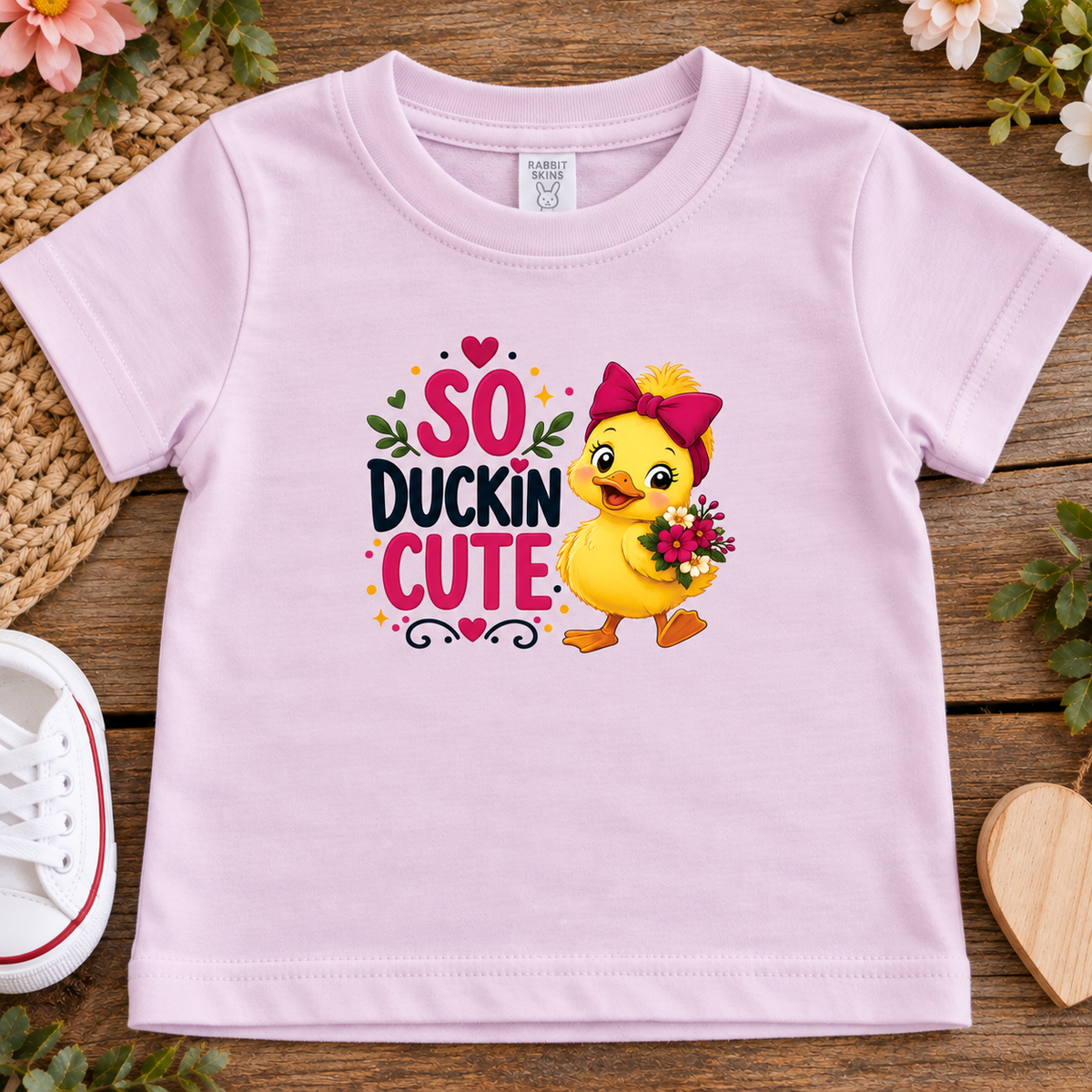 A ballerina pink short-sleeve toddler t-shirt laid flat on a rustic wooden surface, featuring a 'SO DUCKIN' CUTE' graphic with a yellow duckling holding flowers.