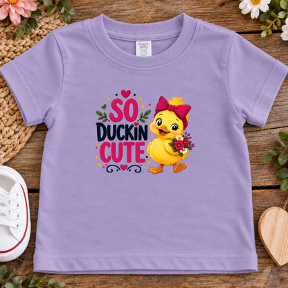 A lavender purple short-sleeve t-shirt for toddler, featuring a colorful graphic print of a yellow duckling holding flowers and the text 'SO DUCKIN CUTE'. The t-shirt is displayed in a flat-lay arrangement on a rustic wooden surface with decorative flowers, a woven mat, and a white sneaker.