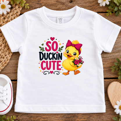 A white short-sleeve toddler t-shirt laid flat on a rustic wooden surface, featuring a colorful graphic of a cute yellow duckling with a pink bow and the text 'SO DUCKIN CUTE'. The shirt is styled with a woven mat, white flowers, a white sneaker, and a wooden heart.