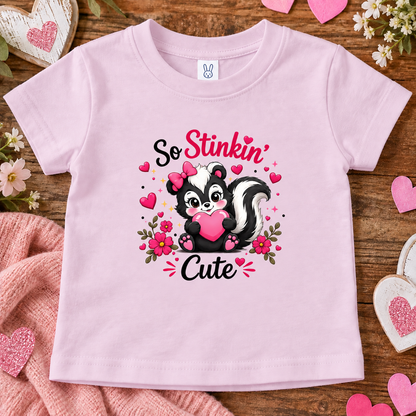 A ballerina pink short-sleeve baby t-shirt with a cute skunk graphic holding a pink heart, surrounded by the words 'So Stinkin' Cute' in black and pink lettering. The t-shirt is displayed in a flat-lay arrangement on a rustic wooden surface with pink hearts, a pink knit blanket, and small white flowers.
