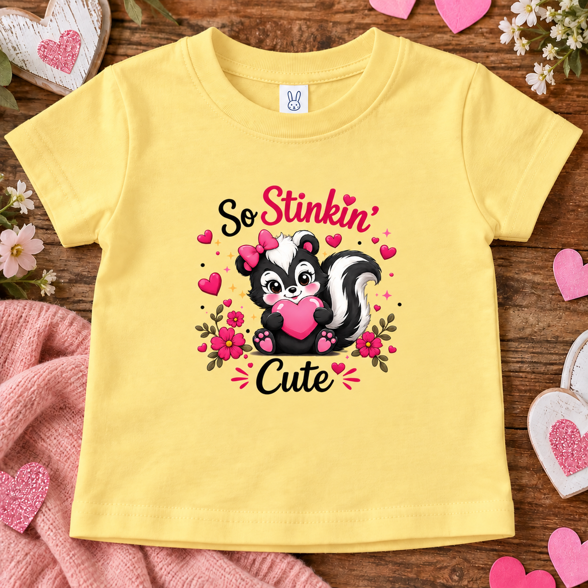 A butter yellow short-sleeve baby t-shirt with a cute cartoon skunk graphic and the text 'So Stinkin' Cute'. The shirt is displayed in a flat-lay arrangement on a rustic wooden background with pink hearts, a pink knit blanket, and small white flowers.