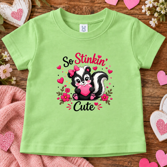 A keylime green short-sleeve kids' t-shirt with a cute cartoon skunk graphic holding a pink heart, surrounded by hearts and the words 'So Stinkin' Cute'. The shirt is displayed in a flat-lay arrangement on a wooden surface with pink hearts, flowers, and a pink blanket.