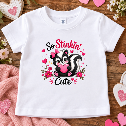 A white short-sleeve baby t-shirt with a cute skunk graphic that reads 'So Stinkin' Cute' in pink and black lettering, surrounded by pink hearts and flowers. The shirt is displayed in a flat-lay arrangement on a wooden surface with pink hearts, a pink knit blanket, and small white flowers.
