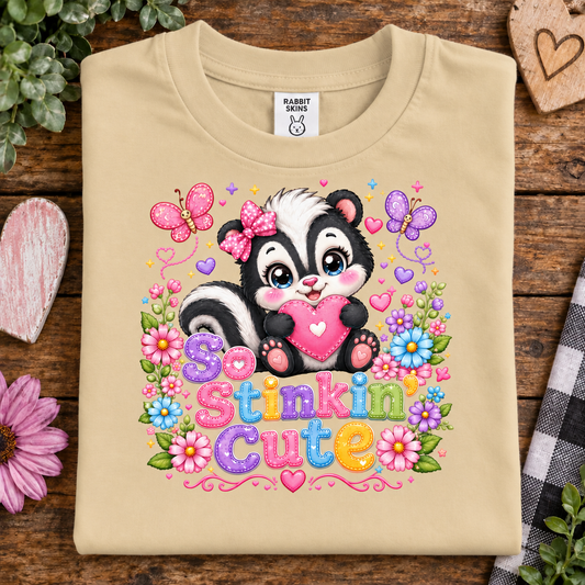 A latte colored crewneck t-shirt with a colorful graphic of a cute cartoon skunk holding a pink heart, surrounded by flowers, butterflies, and the text 'So St nkin Cute'. The shirt is displayed flat on a rustic wooden background with decorative props.
