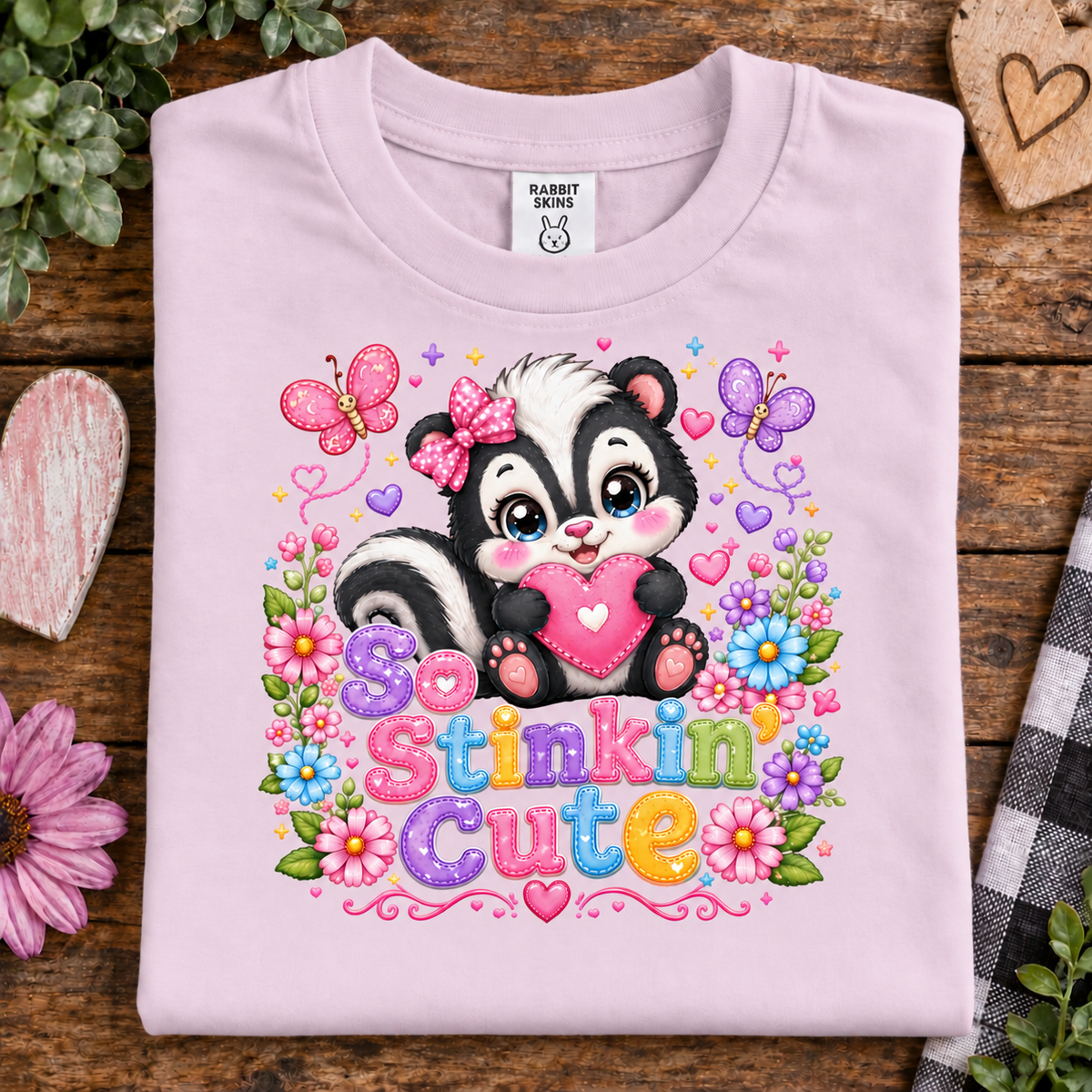 A ballerina pink folded t-shirt with a colorful graphic of a cartoon skunk holding a pink heart, surrounded by flowers, butterflies, and the text 'So Stinkin Cute'. The shirt is displayed on a rustic wooden background with decorative props.