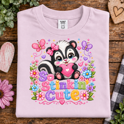 A ballerina pink folded t-shirt with a colorful graphic of a cartoon skunk holding a pink heart, surrounded by flowers, butterflies, and the text 'So Stinkin Cute'. The shirt is displayed on a rustic wooden background with decorative props.