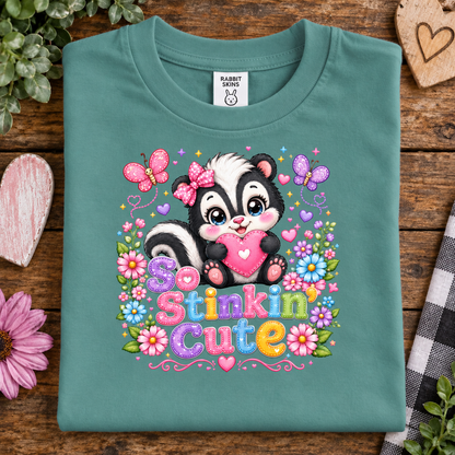 A saltwater (teal) folded t-shirt with a colorful cartoon skunk graphic and the text 'So Stinkin' Cute' on a rustic wooden background with decorative flowers and a heart-shaped sign.