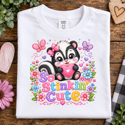 A white short-sleeve t-shirt with a colorful graphic of a cartoon skunk holding a pink heart, surrounded by flowers, butterflies, and the text 'So Stinkin Cute'. The shirt is displayed flat on a rustic wooden surface with decorative props.