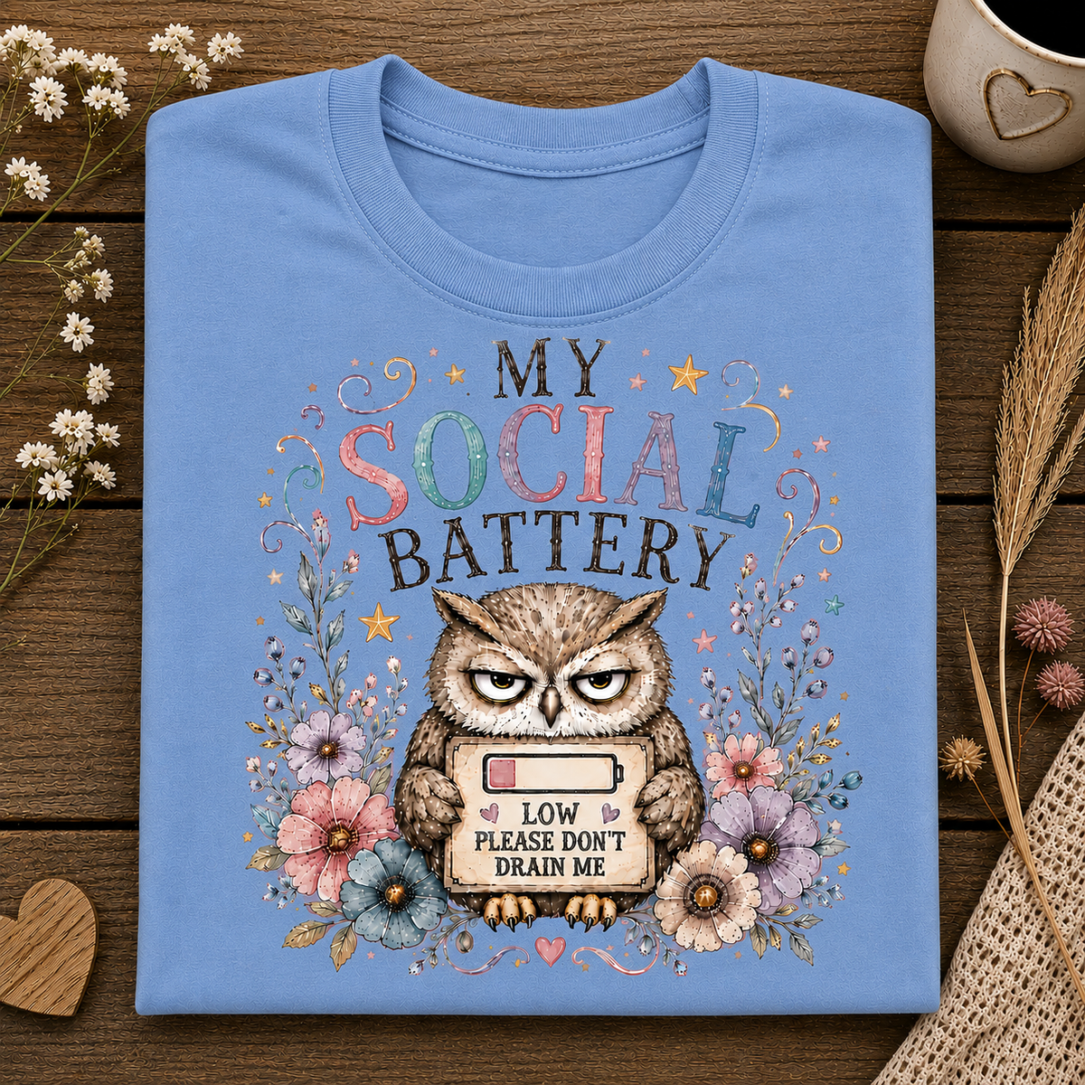 A carolina blue crewneck t-shirt laid flat on a dark wood background, featuring a graphic of an owl holding a low battery symbol with the text 'MY SOCIAL BATTERY LOW PLEASE DON'T DRAIN ME' surrounded by flowers.