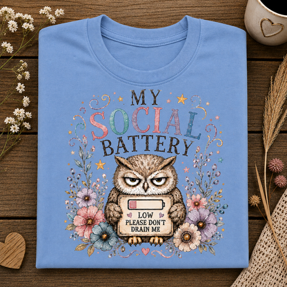 A carolina blue crewneck t-shirt laid flat on a dark wood background, featuring a graphic of an owl holding a low battery symbol with the text 'MY SOCIAL BATTERY LOW PLEASE DON'T DRAIN ME' surrounded by flowers.