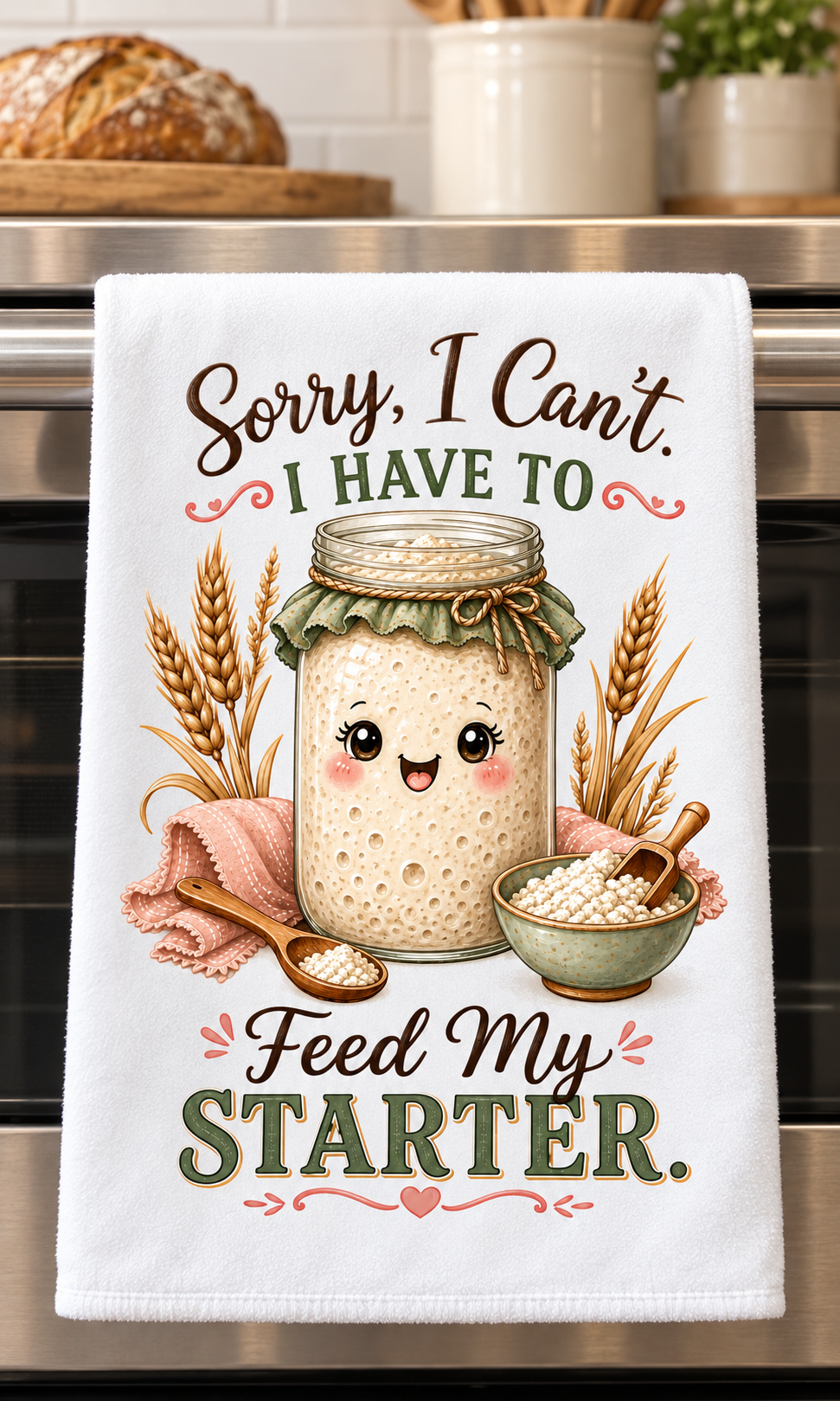 A white kitchen towel hanging on a stainless steel oven handle, featuring a cute cartoon illustration of a smiling sourdough starter jar surrounded by wheat stalks, a bowl of flour, and the text 'Sorry, I Can't. I HAVE TO Feed My STARTER.