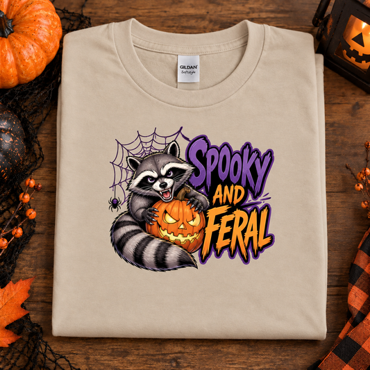 A folded sand colored Gildan t-shirt with a 'Spooky and Feral' raccoon graphic, displayed on a rustic wooden background with Halloween props like a pumpkin, spiderwebs, and a lantern.
