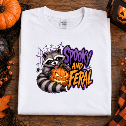 A folded white Gildan Straightweight t-shirt with a colorful 'Spooky and Feral' graphic featuring a raccoon and a jack-o'-lantern, styled on a rustic wooden background with Halloween props.