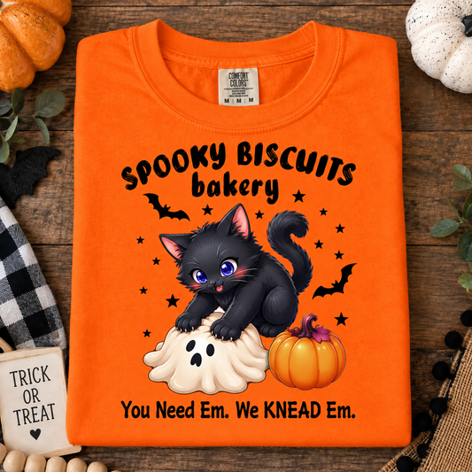 An orange folded t-shirt with a Halloween graphic design featuring a black kitten on a ghost and a pumpkin, with the text 'Spooky Biscuits bakery' and 'You Need Em. We KNEAD Em.