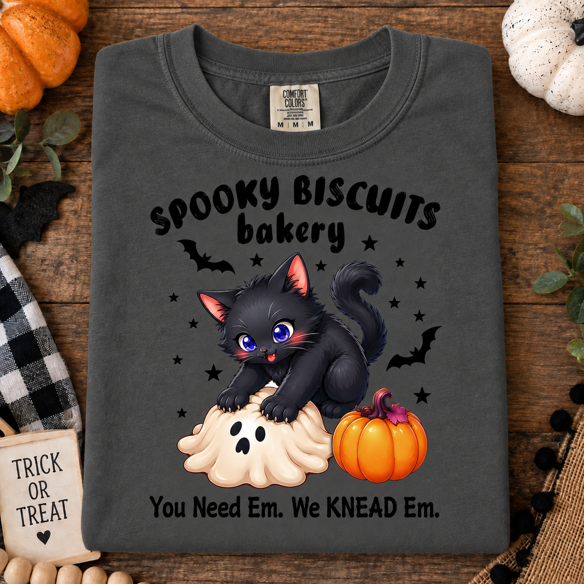 A granite graphic t-shirt laid flat on a rustic wooden surface with Halloween props. The shirt features a cartoon black cat with purple eyes standing on a ghost biscuit, surrounded by black bats and stars, with the text 'SPOOKY BISCUITS bakery' and 'You Need Em.