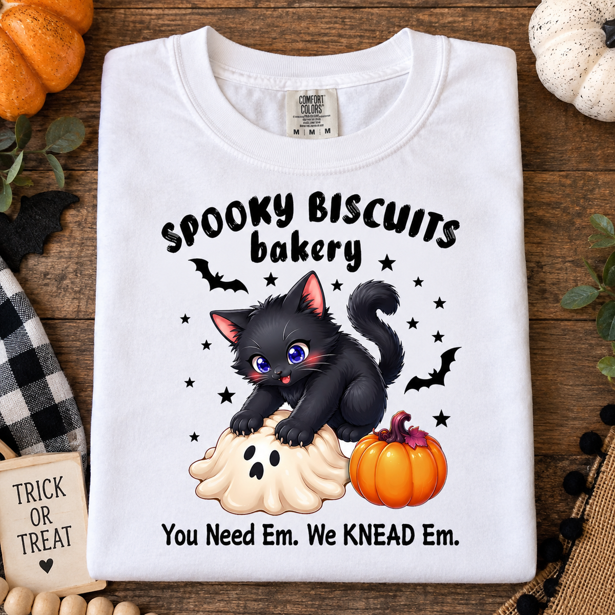 A folded white graphic T-shirt displayed on a rustic wooden surface with Halloween-themed props. The shirt features a black kitten sitting on a ghost cookie, with the text 'Spooky Biscuits Bakery' and 'You Need Em.