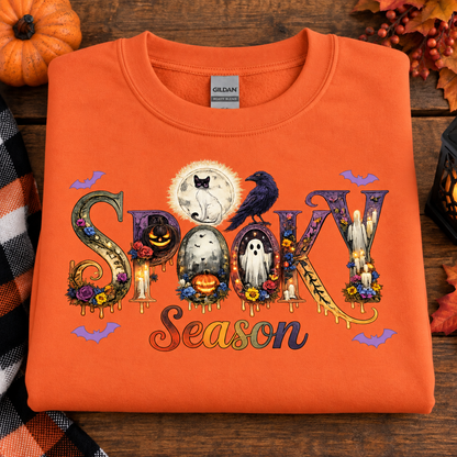 An orange folded crewneck sweatshirt with a colorful 'Spooky Season' graphic, displayed on a rustic wooden background with autumn decorations.