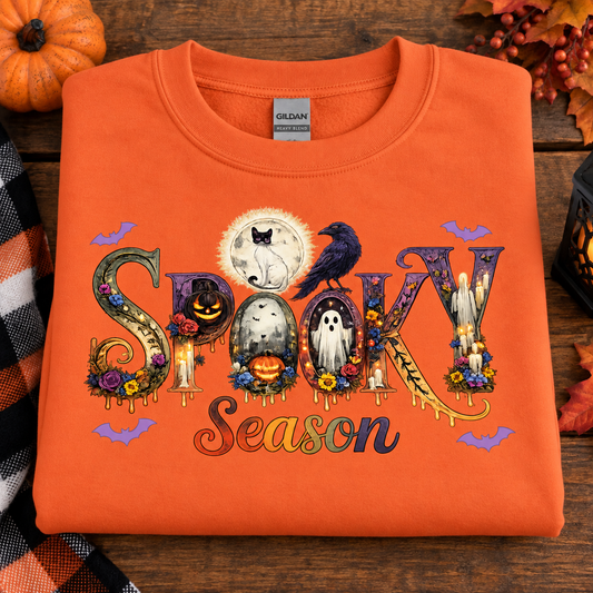 An orange folded crewneck sweatshirt with a colorful 'Spooky Season' graphic, displayed on a rustic wooden background with autumn decorations.