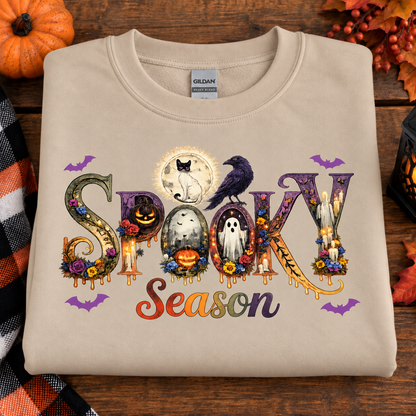 A folded beige Gildan sweatshirt with a colorful 'Spooky Season' graphic, displayed on a rustic wooden background with autumn decorations.