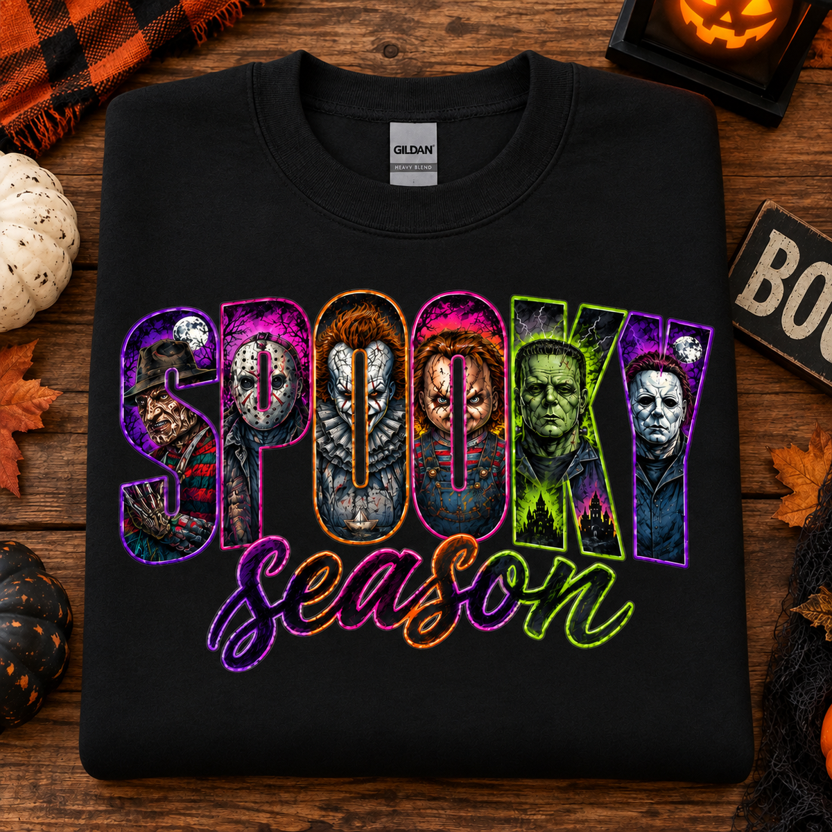 A black Gildan t-shirt laid flat on a rustic wooden surface, decorated with Halloween props like a pumpkin, spiderweb, and autumn leaves. The shirt features a colorful graphic that reads 'SPOOKY Season' with horror movie character illustrations inside the letters.