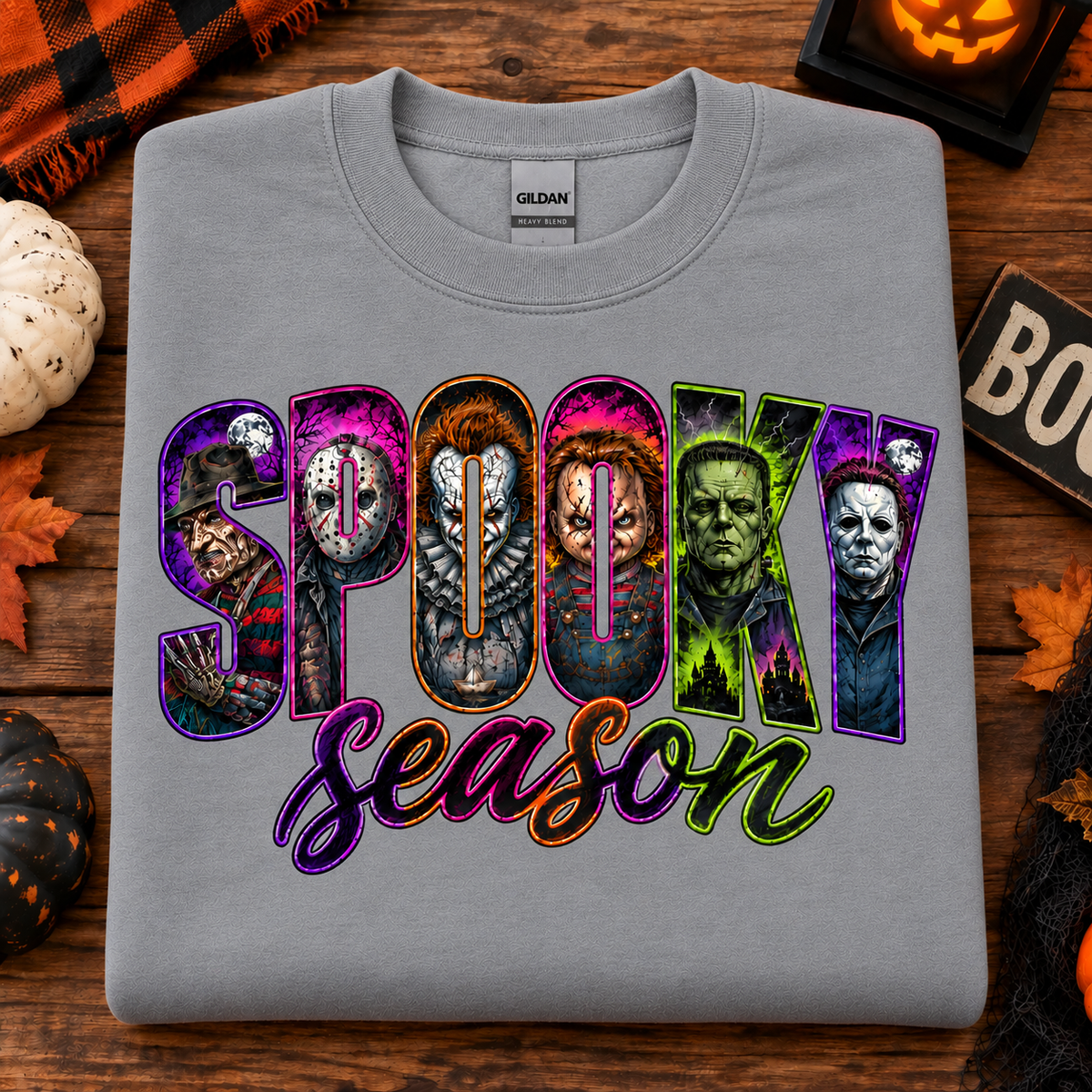 A folded sport gray Gildan t-shirt with a 'Spooky Season' graphic featuring horror movie characters, displayed on a rustic wooden background with Halloween decorations.