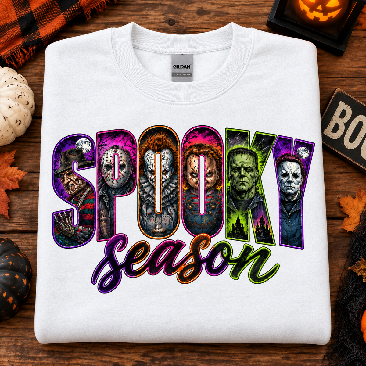 A white Gildan t-shirt laid flat on a rustic wooden surface, decorated with Halloween props like a pumpkin, spiderwebs, and autumn leaves. The shirt features a colorful graphic print that reads 'Spooky Season' with horror movie character silhouettes inside the letters.