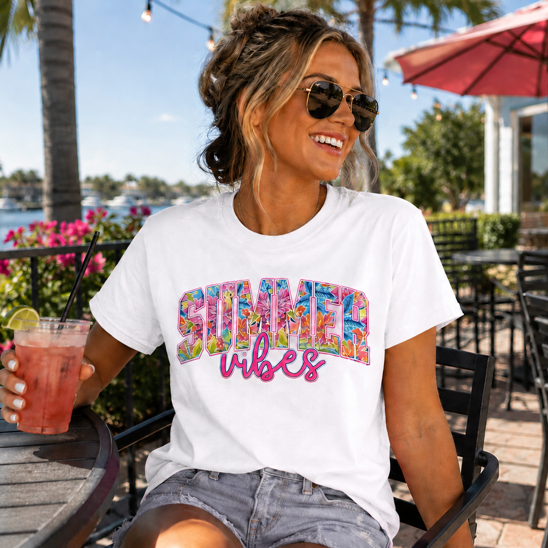 Woman wearing bright colored 'Summer Vibes' white t-shirt holding a drink outdoors.