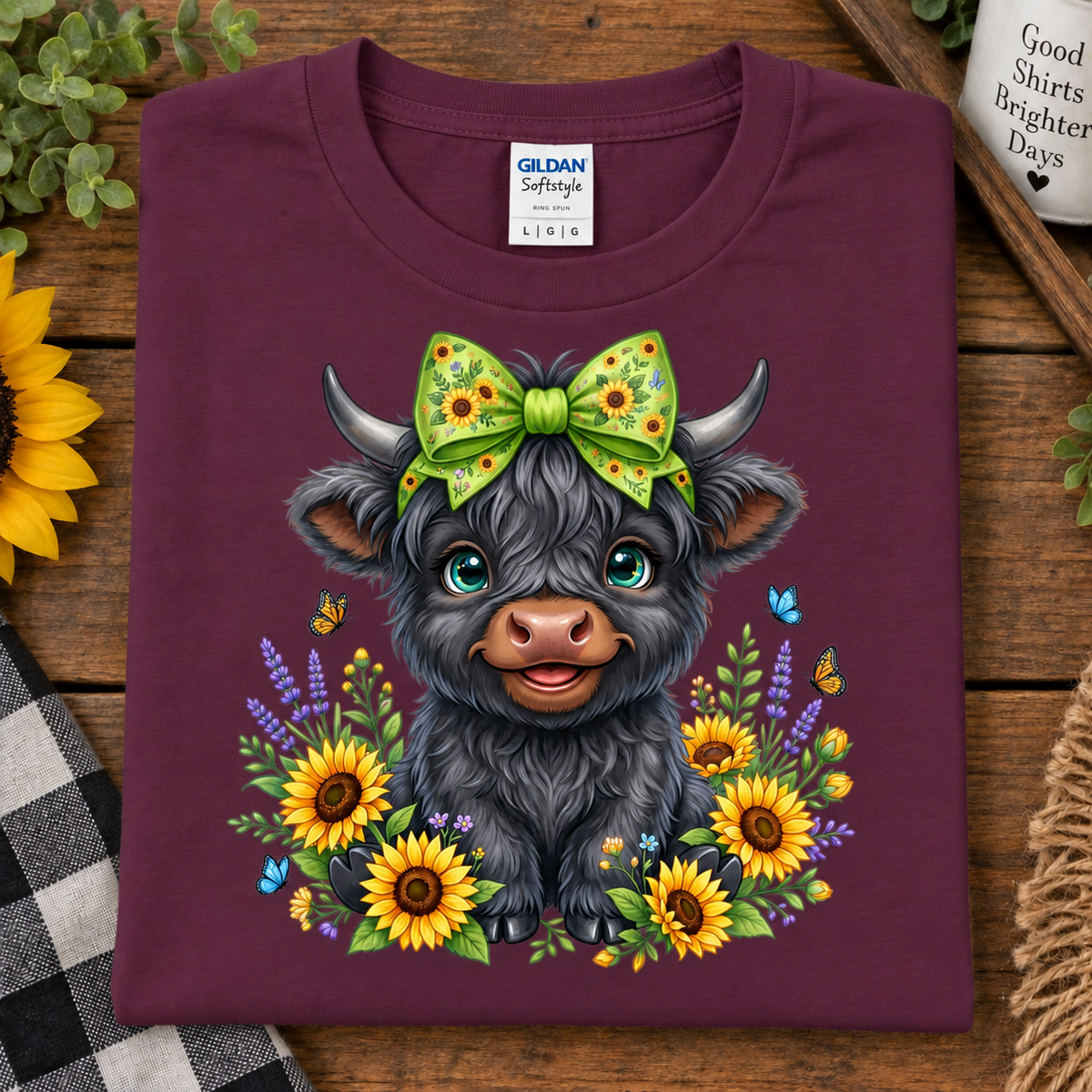 A folded maroon Gildan Softstyle crewneck t-shirt with a colorful graphic of a cute black Scottish Highland cow wearing a green sunflower-patterned bow, surrounded by sunflowers and butterflies, displayed on a rustic wooden background.