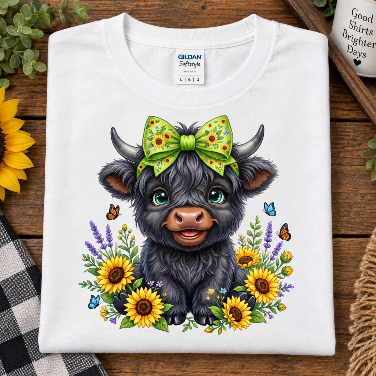 A folded white Gildan Softstyle T-shirt laid flat on a rustic wooden surface, featuring a colorful cartoon illustration of a black Scottish highland cow with a green sunflower-patterned bow, surrounded by yellow sunflowers, purple lavender, blue butterflies, and green foliage.