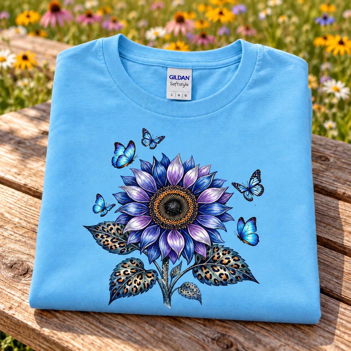 A folded light blue Gildan Softstyle t-shirt is displayed on a wooden bench with a blurred background of a flower field. The shirt features a detailed graphic of a purple and blue sunflower with butterflies and leopard-print leaves.
