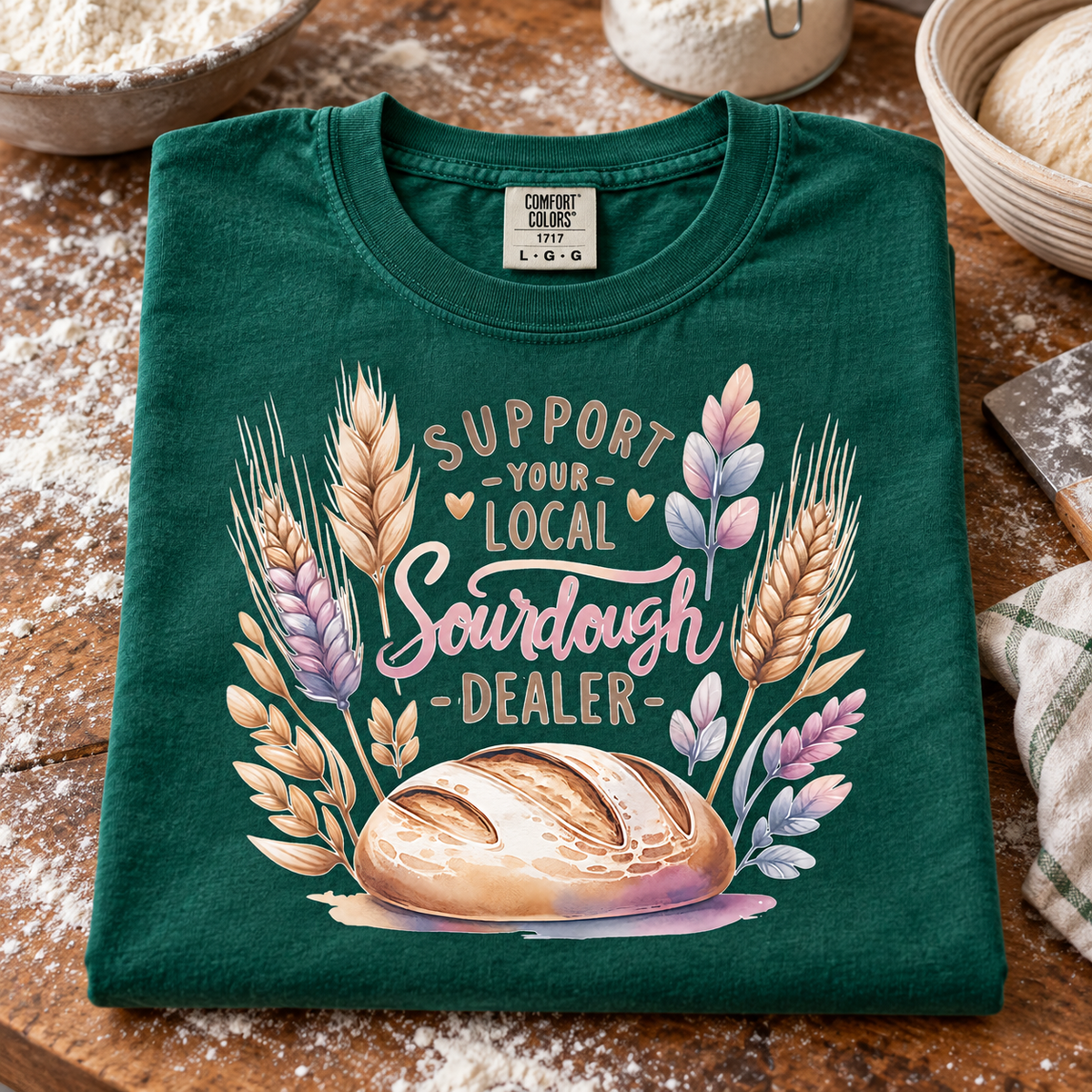 A folded emerald t-shirt with a 'Support Your Local Sourdough Dealer' graphic featuring wheat stalks and a loaf of bread, displayed on a rustic wooden table with flour and baking props.