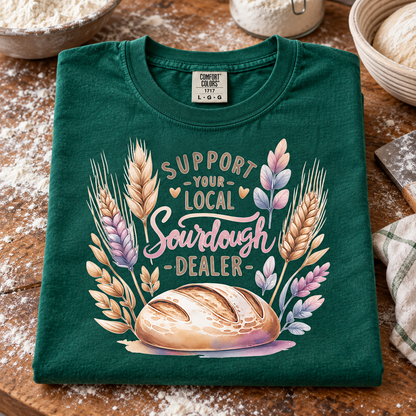 A folded emerald t-shirt with a 'Support Your Local Sourdough Dealer' graphic featuring wheat stalks and a loaf of bread, displayed on a rustic wooden table with flour and baking props.