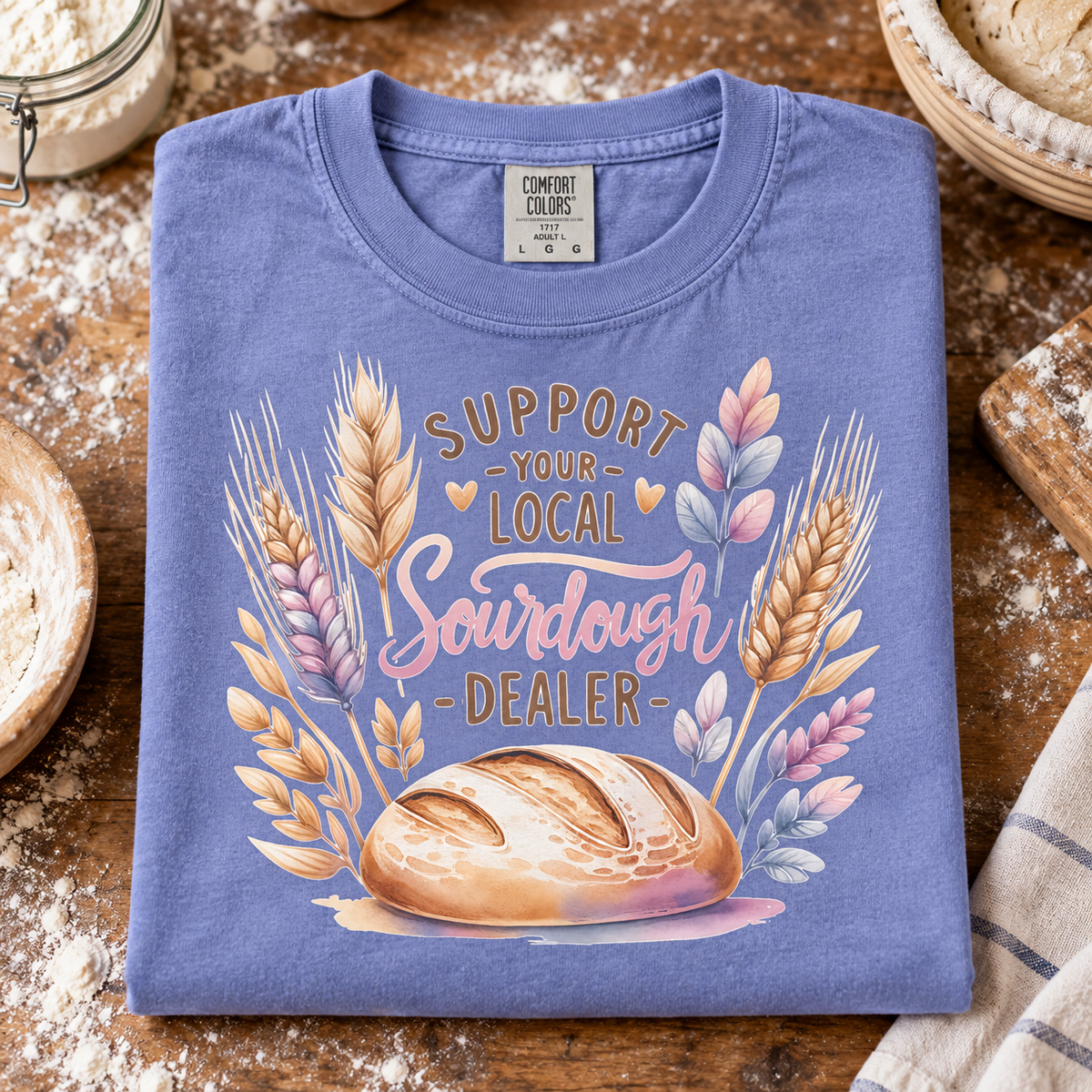 A folded periwinkle blue t-shirt with a watercolor graphic of a sourdough bread loaf and wheat stalks, featuring the text 'SUPPORT YOUR LOCAL SOURDOUGH DEALER'. The shirt is styled on a rustic wooden table surrounded by baking props like flour, a jar, and a cloth.