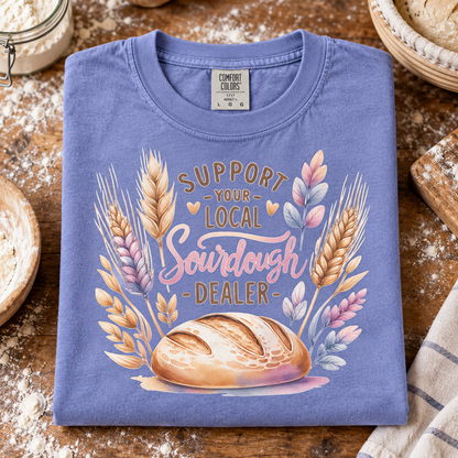 A folded periwinkle blue t-shirt with a watercolor graphic of a sourdough bread loaf and wheat stalks, featuring the text 'SUPPORT YOUR LOCAL SOURDOUGH DEALER'. The shirt is styled on a rustic wooden table surrounded by baking props like flour, a jar, and a cloth.