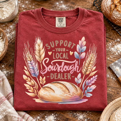 A folded crimson red graphic T-shirt displayed on a rustic wooden table with flour-dusted bread, a cloth, and baking tools around it. The shirt reads “SUPPORT YOUR LOCAL Sourdough DEALER” with wheat stalks and a loaf of bread in a pastel watercolor-style print.