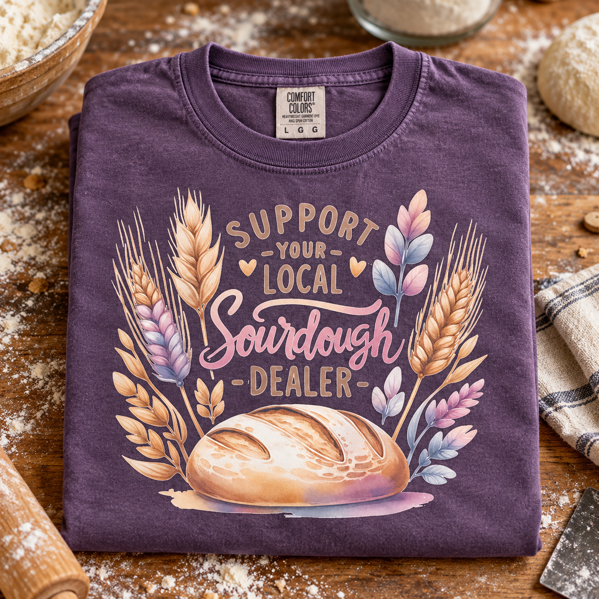 A folded grape purple t-shirt with a watercolor graphic of a sourdough bread loaf and wheat stalks, featuring the text 'SUPPORT YOUR LOCAL Sourdough DEALER'. The shirt is styled on a rustic wooden table with flour, a rolling pin, and baking ingredients.