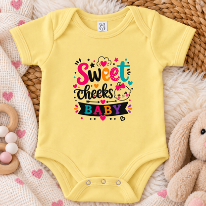 A butter colored graphic onesie design with the words 'Sweet cheeks BABY' in playful hand-lettered typography, surrounded by hearts, stars, dots, and a cartoon baby face with a pink bow on a white background.