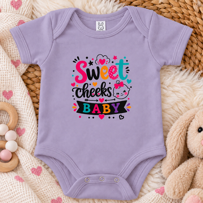 A lavender colored graphic onesie with the words 'Sweet cheeks BABY' in playful hand-lettered typography, surrounded by hearts, stars, dots, and a cartoon baby face with a pink bow on a white background.