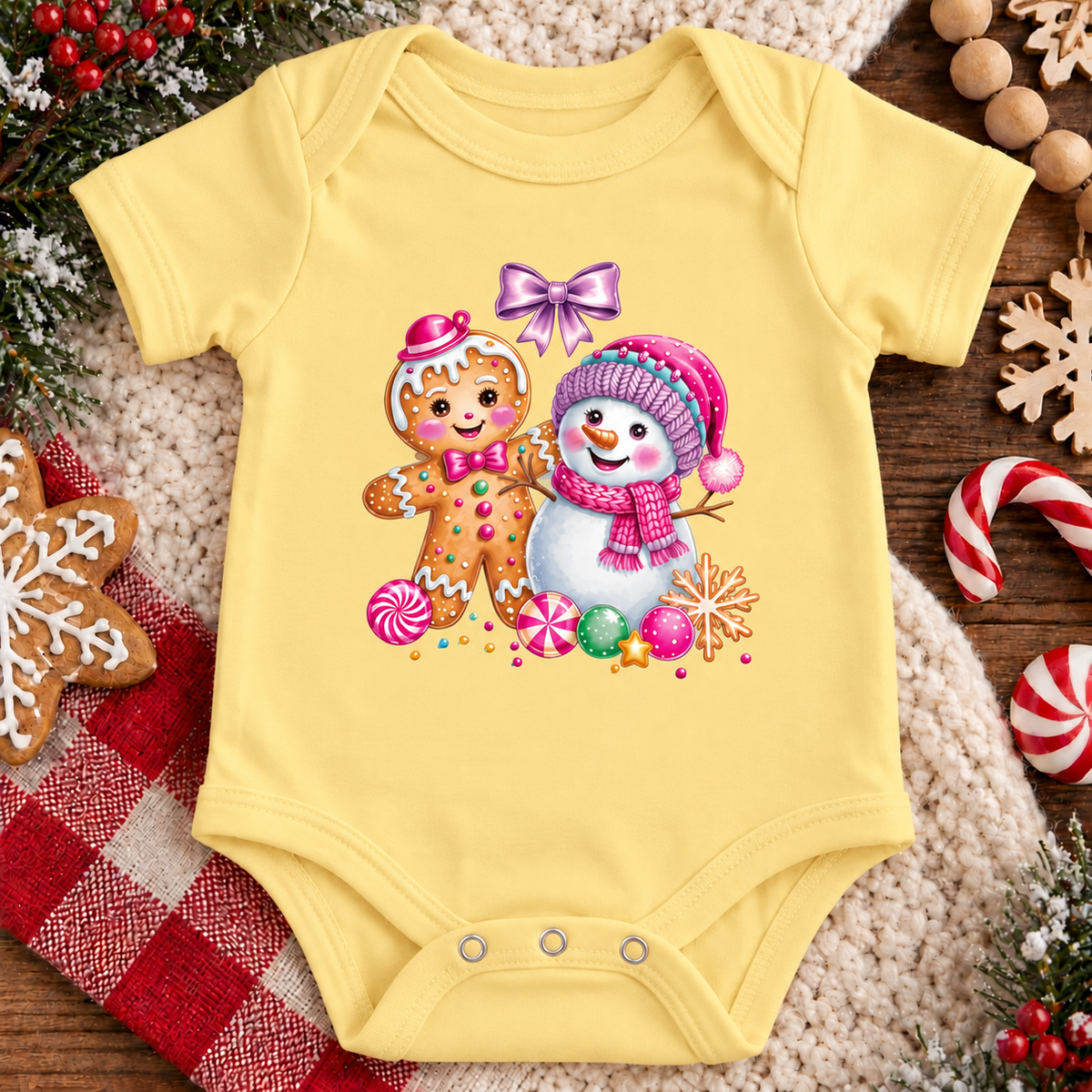 A butter yellow baby bodysuit with a colorful Christmas print of a gingerbread man and a snowman, laid flat on a cozy beige knit blanket surrounded by holiday decorations like candy canes, a gingerbread cookie, and pine branches.