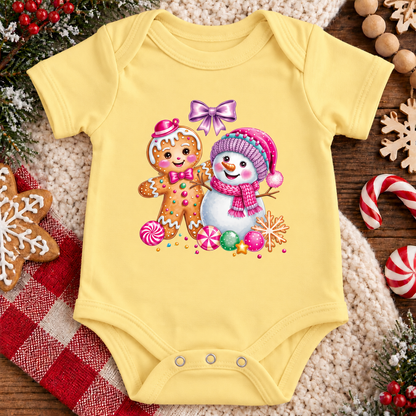 A butter yellow baby bodysuit with a colorful Christmas print of a gingerbread man and a snowman, laid flat on a cozy beige knit blanket surrounded by holiday decorations like candy canes, a gingerbread cookie, and pine branches.