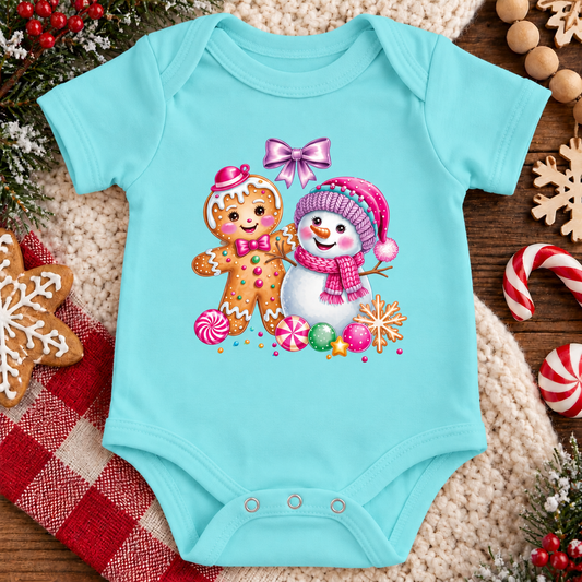 A chill blue baby bodysuit with a colorful Christmas print of a gingerbread man and a snowman, styled on a cozy beige knit blanket with holiday decorations.