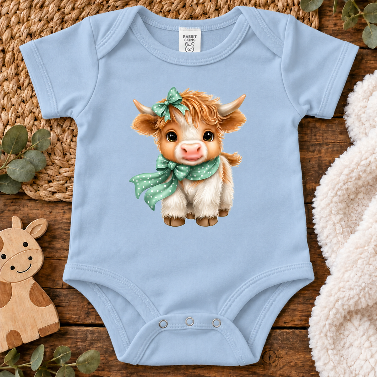 A light blue colored onesie(bodysuit) with a highland cow with a polka dot scarf and bow.