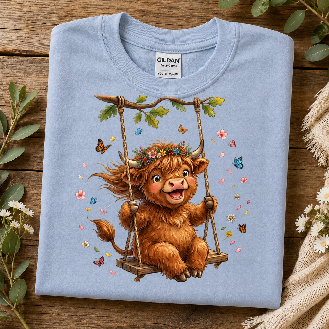 A folded light blue Gildan Heavy Cotton T-shirt is displayed on a rustic wooden surface, decorated with green leaves and white flowers. The shirt features a colorful graphic of a fluffy brown Highland cow sitting on a swing surrounded by butterflies and flowers.