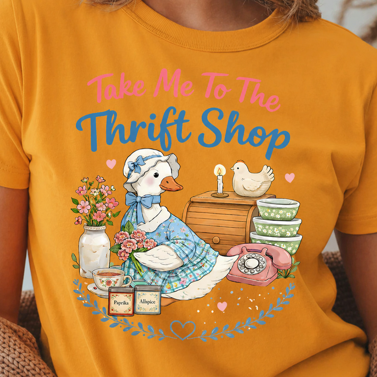 Person wearing a gold colored t-shirt with a graphic of a duck in a thrift store setting and text 'Take Me To The Thrift Shop'.