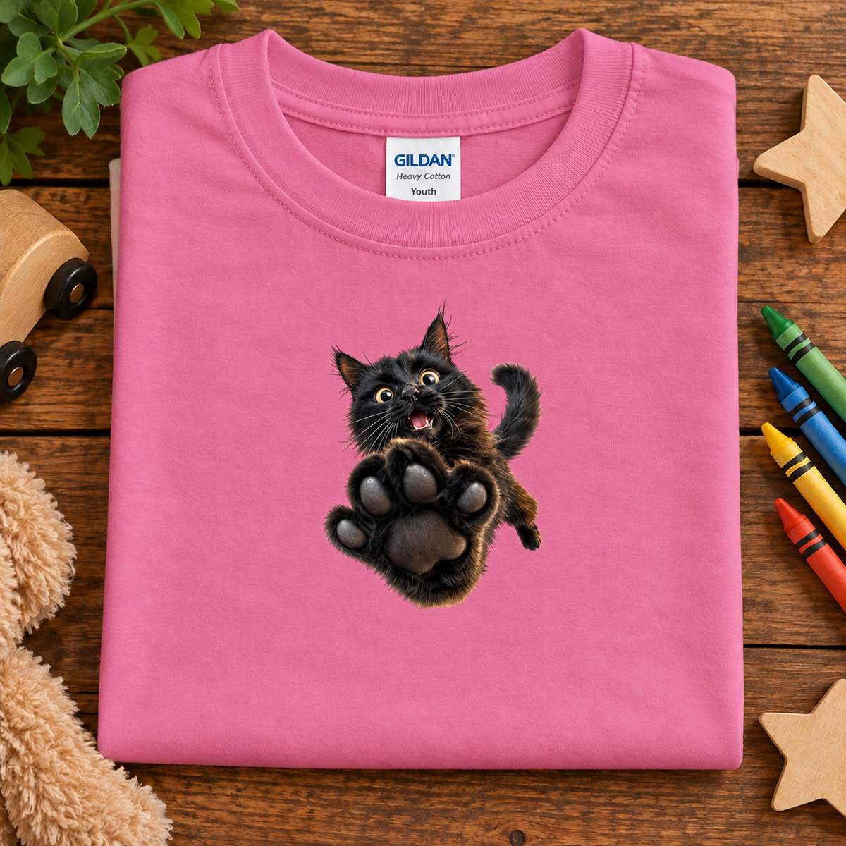 A folded azalea pink Gildan Youth t-shirt with a graphic of a jumping black cat, displayed on a wooden surface with toys and crayons.