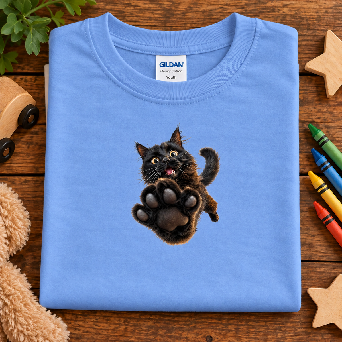 A folded carolina blue Gildan Youth cotton t-shirt with a graphic of a jumping black cat, displayed on a wooden surface with toys and crayons.